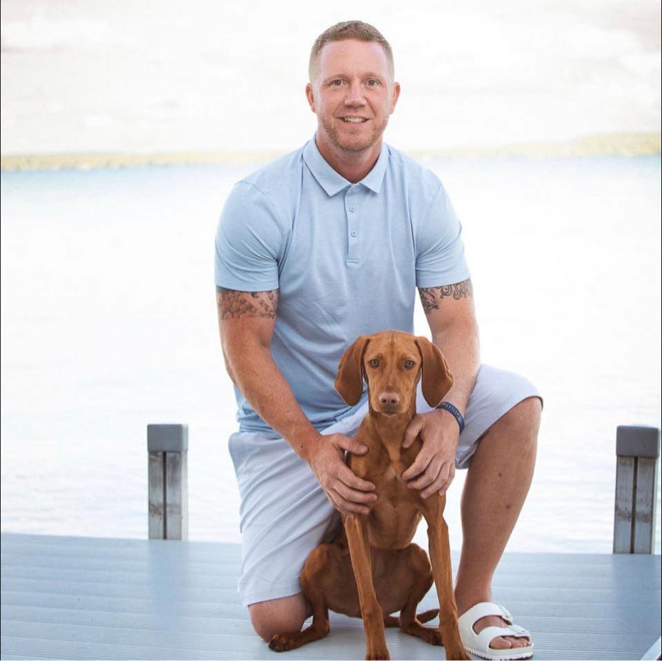 Travis is in a light blue polo shirt and white shorts kneels on a dock, smiling at the camera. He has tattoos on his arms and is accompanied by a brown dog, which he gently holds. The background features a serene lake.